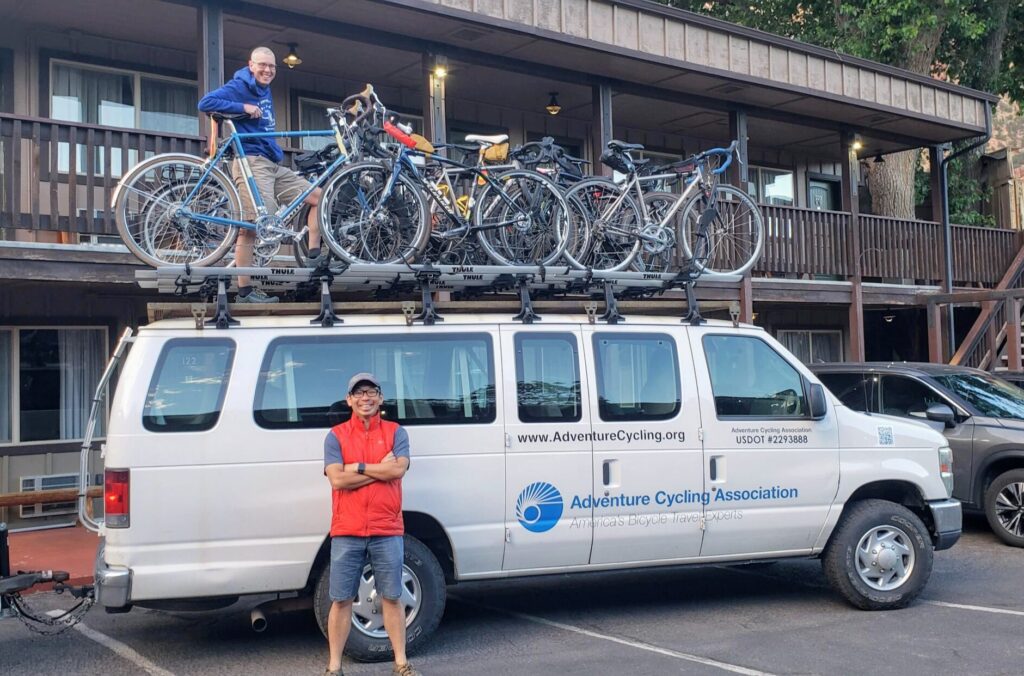 Woman standing on top of white van loaded with many bikes in the parking lot of a two story motel. Man standing in front of van.